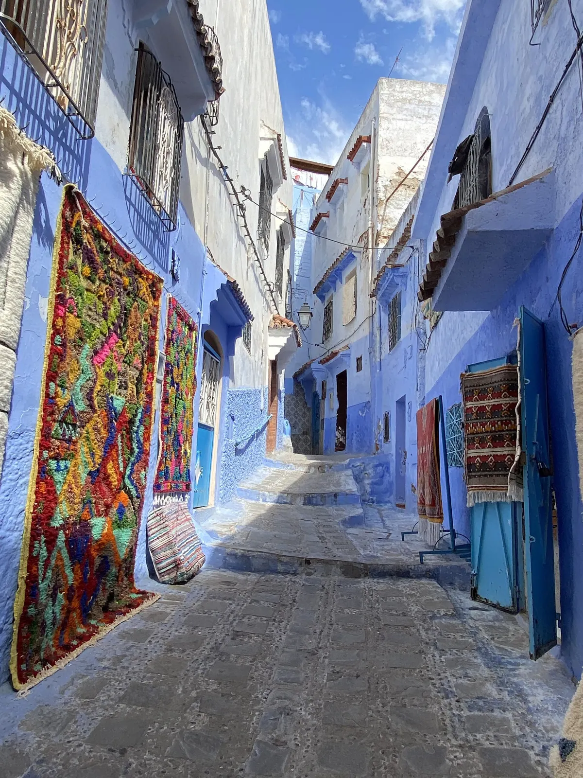 A blue-washed stepped alley in the Chefchaouen medina with a large multicoloured Berber carpet hanging on the left wall, rugs and a striped cushion on the ground beside it, and blue doors opposite