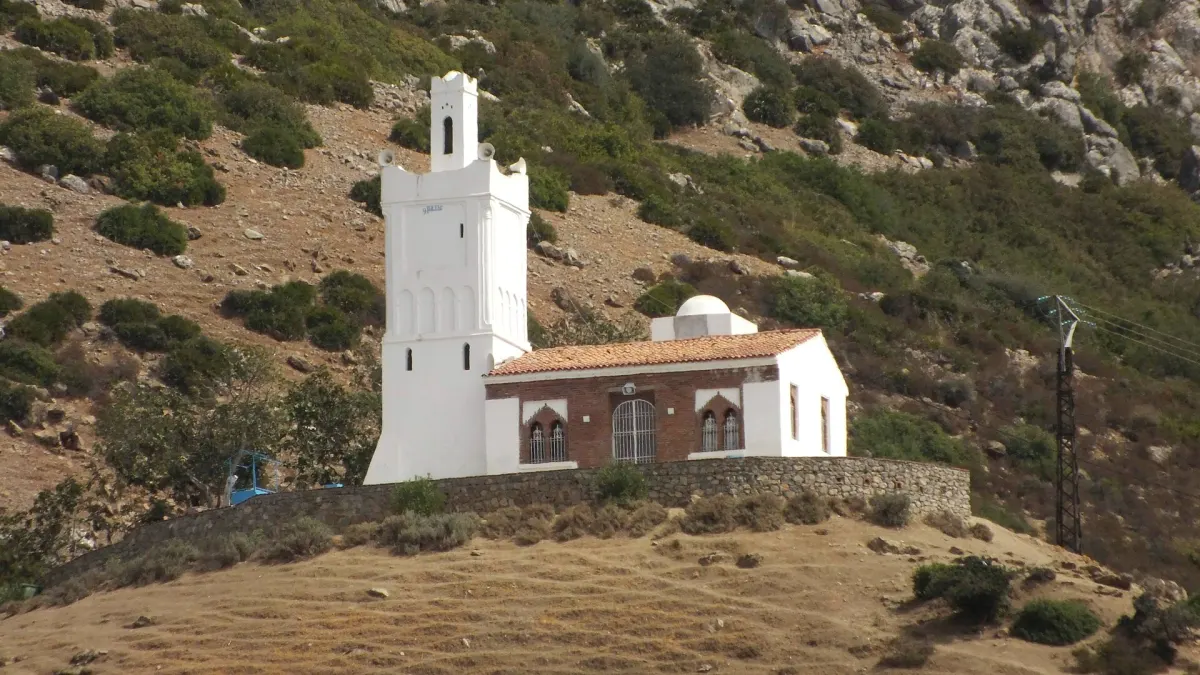 The white Spanish Mosque standing alone on a dry tussocky hillside above Chefchaouen, a square tower with a small dome and horseshoe-arched windows, behind a low stone retaining wall
