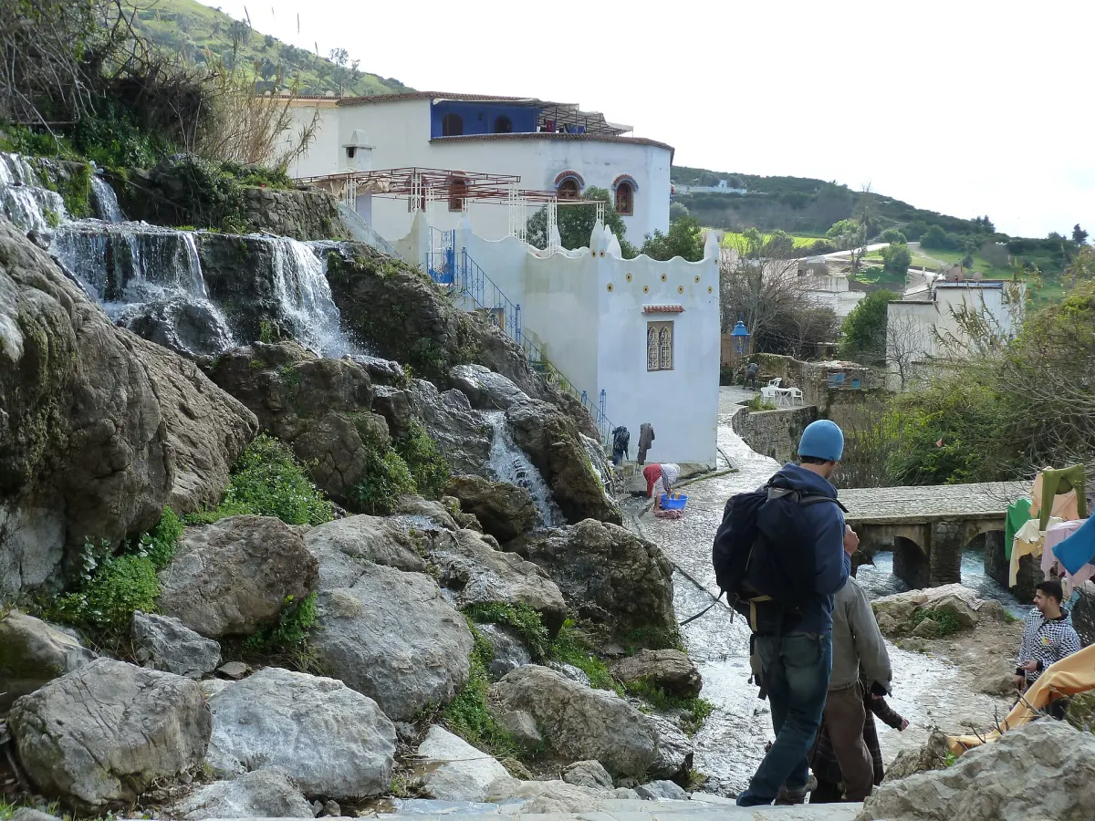 The Ras el Maa stream stepping down over large mossy boulders below white buildings with blue trim, with a walker on a stone path beside the water and a woman washing laundry in a basin at the water's edge