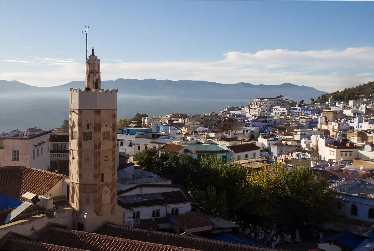The Grand Mosque's brick minaret rising above the blue-and-white rooftops of the Chefchaouen medina, with café tables in a tree-filled square below and the Rif mountains hazy behind