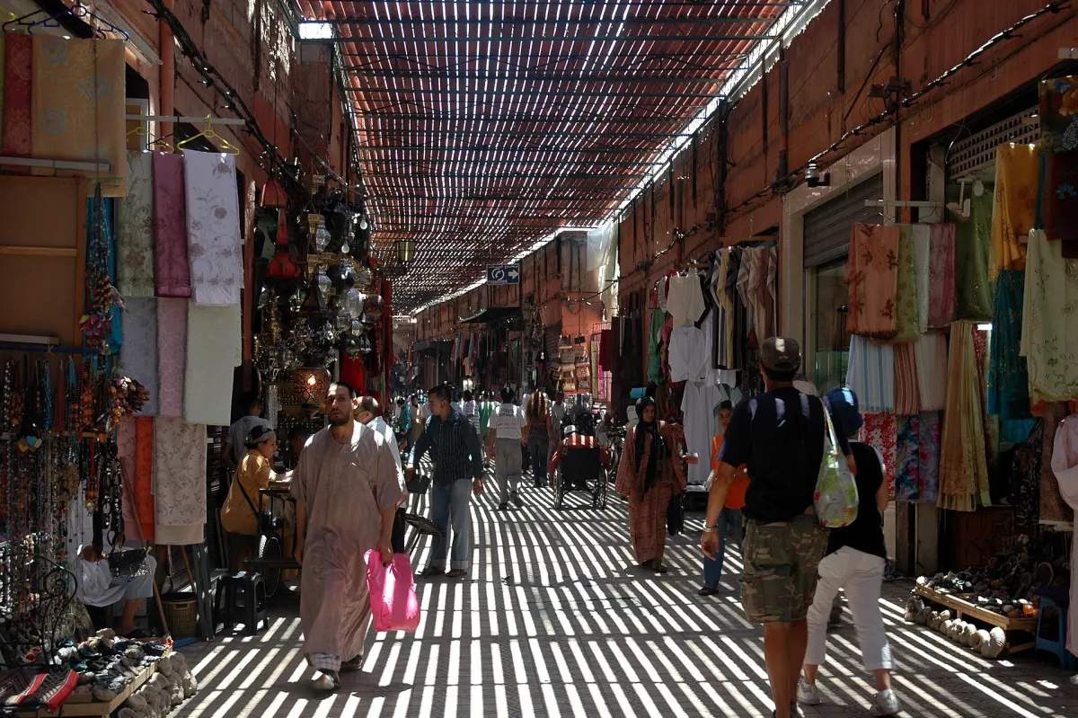 A covered souk alley in the Marrakech medina, striped sunlight through a slatted roof and textiles hanging on both sides