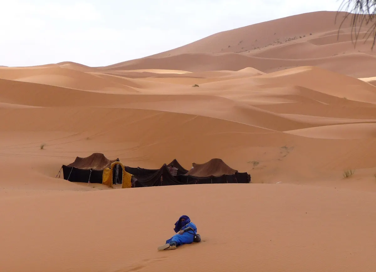 Black goat-hair tents pitched in a hollow between orange dunes, a yellow-fringed opening on one and a person in a blue robe resting on the sand in the foreground