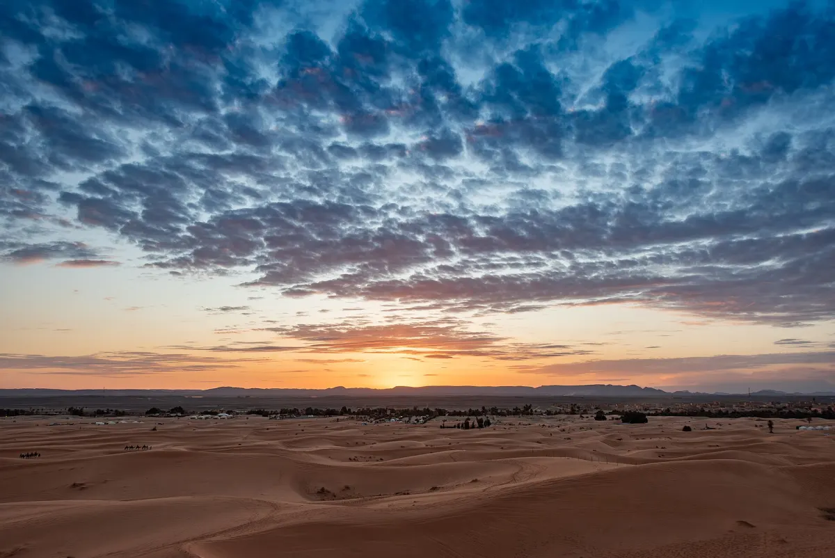 Clouds lit orange by the setting sun over the Erg Chebbi dune field, with the low buildings and palms of the village strung along the horizon at the foot of the dunes